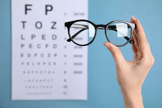 Woman holding glasses against eye chart on blue background, closeup. Ophthalmologist prescription